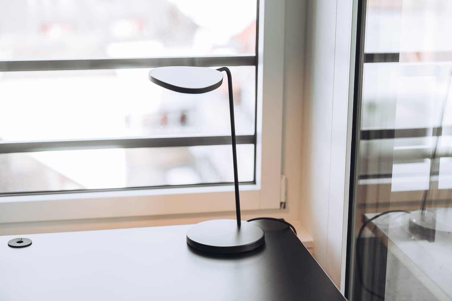 Auf einem Tisch steht eine schlichte schwarze Tischlampe, welche f&uuml;r zus&auml;tzliches Licht sorgen kann